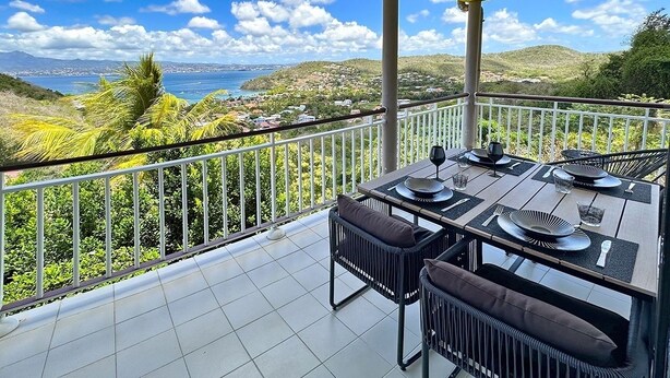 A balcony with a table set for dining, overlooking a coastal landscape with hills and a view of the sea. - Le Loft - Villa Ansalane (Les Trois-Îlets)