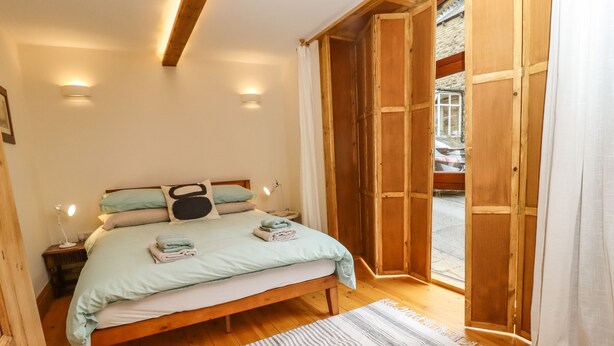 A bedroom with a bed, bedside lamps, a wooden floor, and a view through an open door. - 1 Chews Cottage (Harrogate)