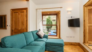 A teal sofa with a matching ottoman, a wooden floor, a window with a view of a parked car and greenery, and a mounted TV. - 1 Chews Cottage (Harrogate)