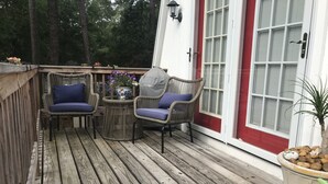 Una terraza de madera con sillas de mimbre, una mesa pequeña, una planta en maceta y una puerta roja.