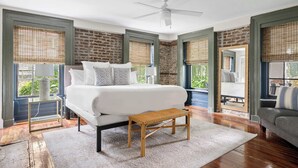 A bedroom with a large bed, a wooden bench, a ceiling fan, and large windows with blinds.