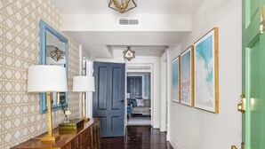 A hallway with patterned wallpaper, a wooden dresser with a lamp, a blue door, and framed artwork on the walls.