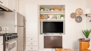 Una cocina moderna con horno y estufa integrados, un armario blanco con cajones, un televisor montado en la pared y una zona de estar con un sofá marrón y una alfombra tejida.