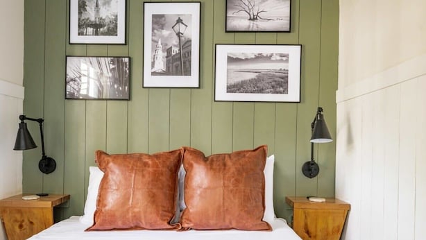Un dormitorio con pared verde, una cama con ropa de cama blanca, dos almohadillas de cuero marrón y cuatro cuadros enmarcados en la pared.