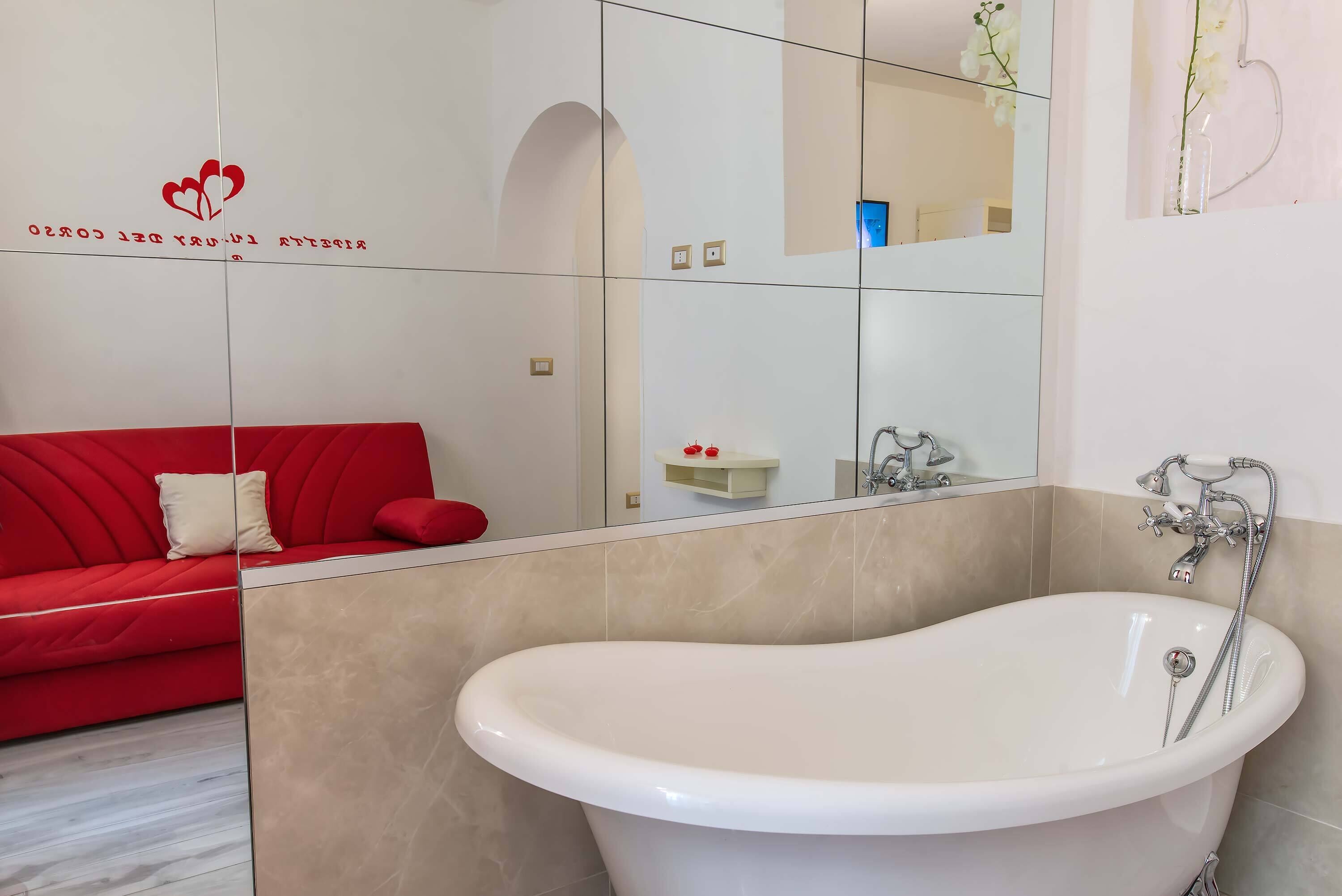 A modern bathroom with a freestanding bathtub, a red sofa with a cushion, and a mirror reflecting the room.