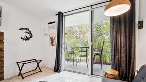 A modern living room with a glass table, a sofa, a wooden side table, and a view of an outdoor patio with chairs and plants.