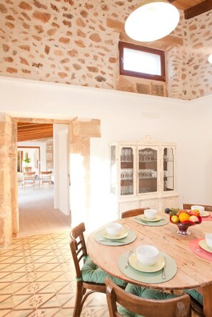 Un coin repas agrémenté d’un mur en pierre, d’un plafond en bois et d’une table à manger mise avec des assiettes, des bols et un saladier de fruits.