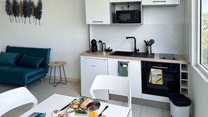 A compact kitchen with a microwave, sink, and oven, a dining table with breakfast items, a teal sofa, and a small side table. - Le Turquoise (Les Trois-Îlets)