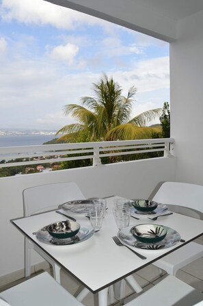 A dining table set with plates, glasses, and cutlery on a balcony overlooking a coastal view with palm trees and a distant cityscape. - Le Turquoise (Les Trois-Îlets)