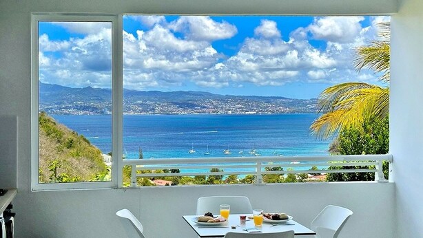 A balcony with a table set for breakfast, overlooking a coastal view with mountains and the sea. - Le Turquoise (Les Trois-Îlets)