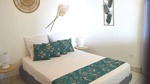 A neatly made bed with a white and green floral bedspread, a small bedside table with a lamp, and decorative wall art including a fan and a feather. - Le Turquoise (Les Trois-Îlets)
