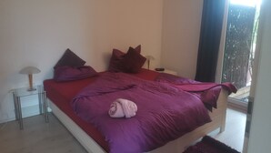 Un dormitorio con cama, almohadas, mesita de noche, lámpara y ventana con cortinas.