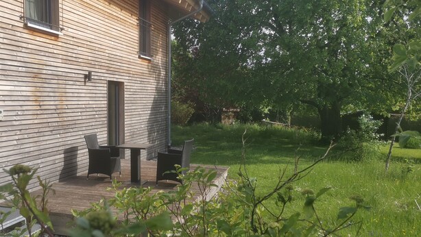 Una casa de madera con puerta y ventanas, un pequeño espacio exterior con mesa y sillas, y un patio con césped y árboles.