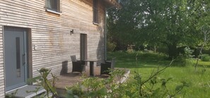 Et træhus med dør og vinduer, et lille udendørs siddeområde med bord og stole, og en græsplæne med træer.