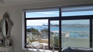 Ein Zimmer mit großem Fenster und Blick auf einen Hafen, ein weißer Stuhl, ein kleiner Tisch und ein dekorativer Spiegel an der Wand.