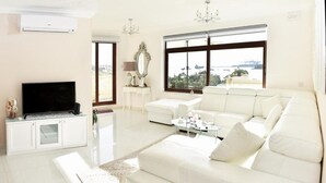 Ein geräumiges Wohnzimmer mit einem weißen Sofa, einem Flachbildfernseher und großen Fenstern mit Blick auf das Meer.