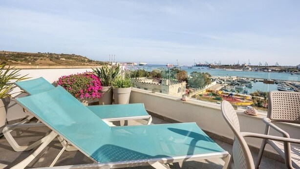 Ein Balkon mit zwei Lounge-Stühlen und einem Tisch, mit Blick auf einen Hafen voller Boote und einer fernen Küste.