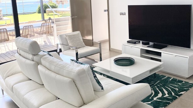 Un salon moderne avec un canapé blanc, une table basse et un téléviseur à écran plat. Une porte-fenêtre coulissante donne accès à un balcon offrant une vue sur l’océan.