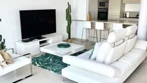 Un salon moderne avec un canapé blanc, un téléviseur à écran plat, un cactus et une cuisine en arrière-plan.