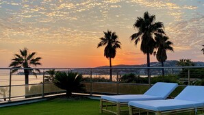 Une terrasse avec deux chaises longues, des palmiers et une vue sur le coucher de soleil au-dessus d’une étendue d’eau.