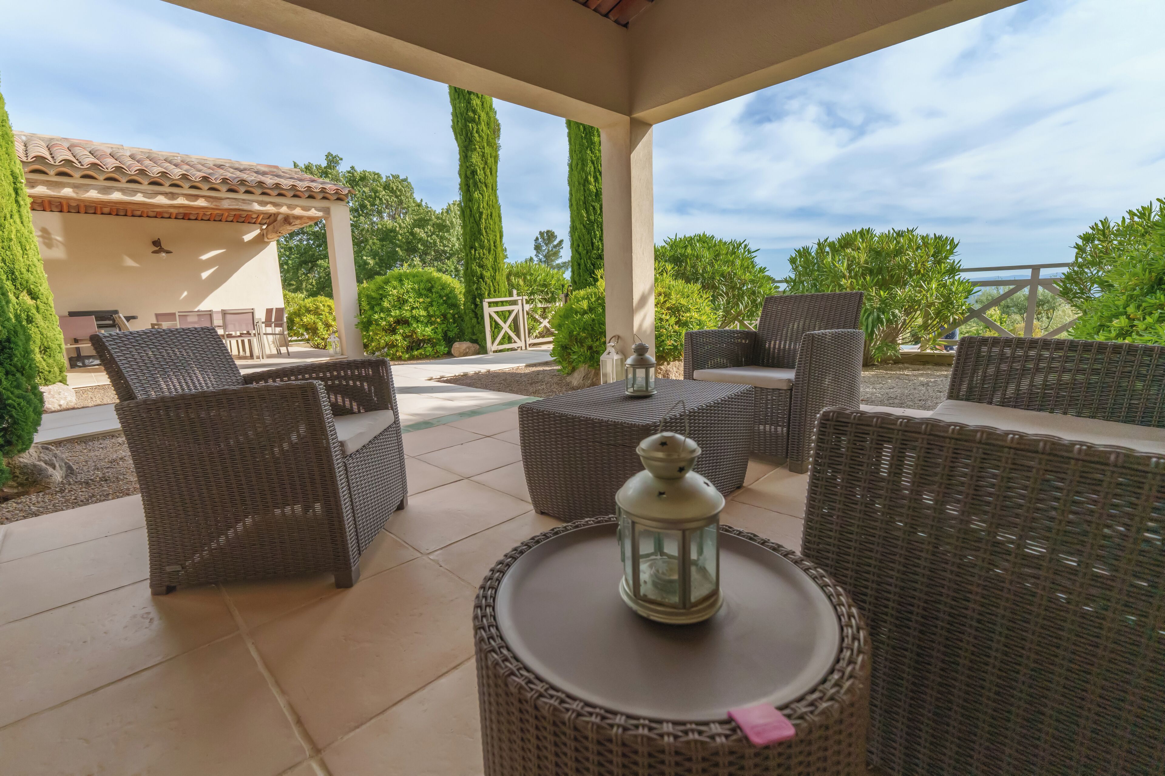 Une terrasse extérieure aménagée avec des meubles en osier, une lanterne et offrant une vue sur le jardin et le ciel environnants.