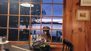 En hund sidder ved et vindue med udsigt til en sø og bjerge i solnedgang.