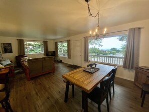 A spacious living room with a wooden dining table, a sofa, and a TV. Large windows offer a view of the outdoors. - The Peaceful Pelican is close to town and just a 5 min walk to the beach! (Brookings)