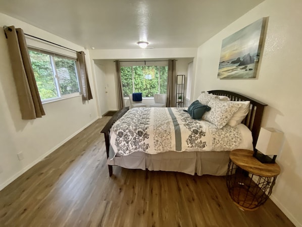 A bedroom with a large bed, a small table, and a view of greenery outside.