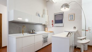 Une cuisine moderne avec des meubles blancs, un four en acier inoxydable, un évier et un micro-ondes encastré. On y trouve également un petit îlot central doté d’un évier et d’un luminaire suspendu.