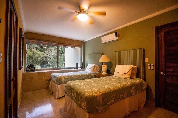 Een kamer met twee bedden, een plafondventilator, een airconditioningsunit en een raam met uitzicht op bomen.