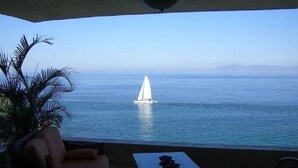 Een balkon met uitzicht op zee, een zeilboot en een palmboom.