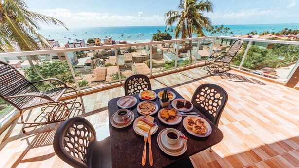 Una mesa de comedor con platos de comida y bebidas, rodeada de sillas, en un balcón con vista al mar y palmeras.
