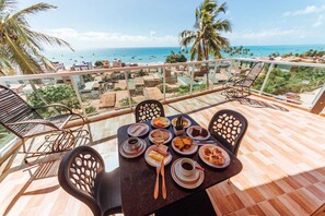 Una mesa de comedor con platos de comida y bebidas, rodeada de sillas, en un balcón con vista al mar y palmeras.