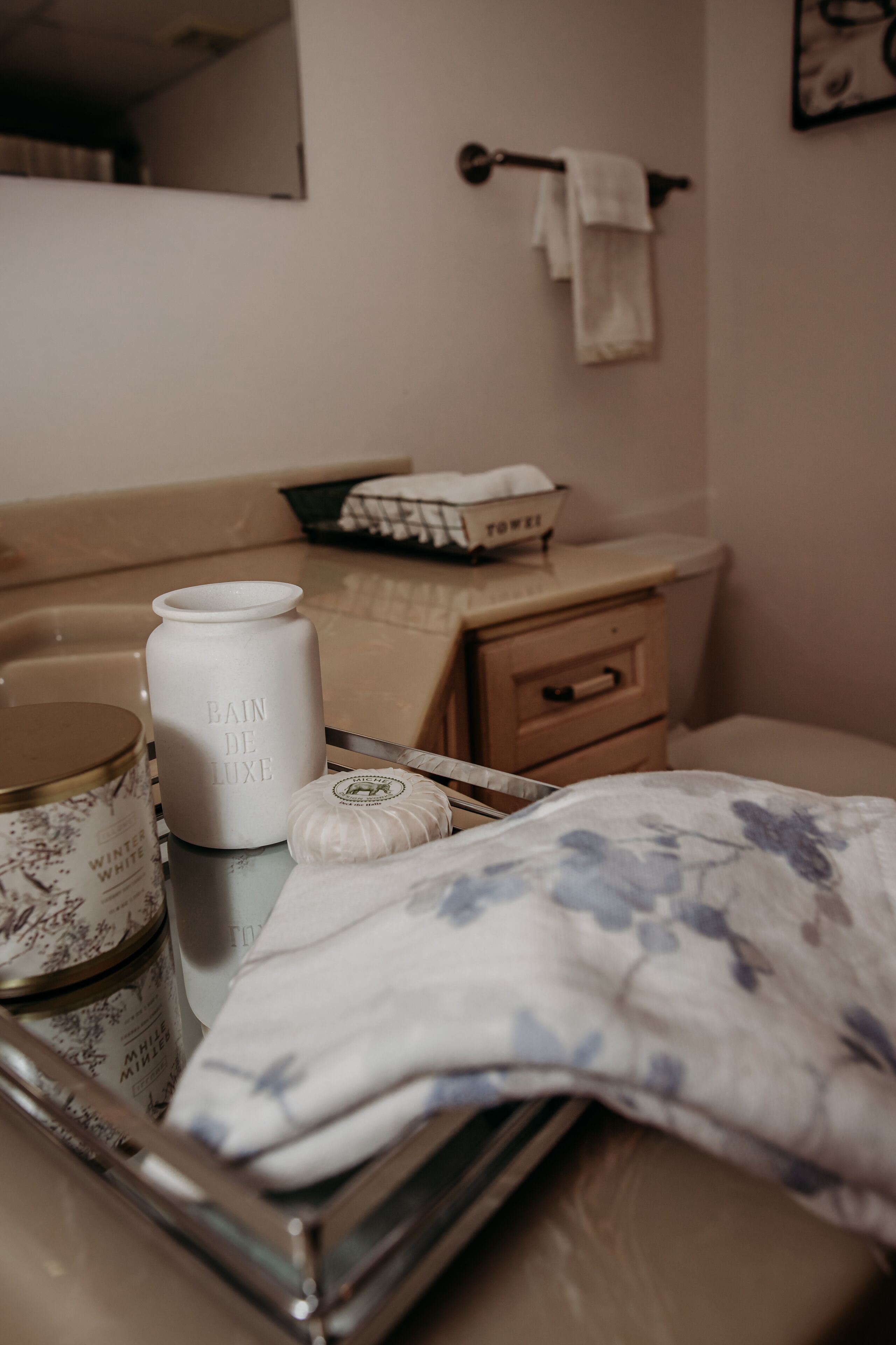 Een badkamer met een beige wastafel, een witte vaas met de tekst 'BAIN DE LUXE', een klein rond potje en een handdoek met bloemenprint op een dienblad.