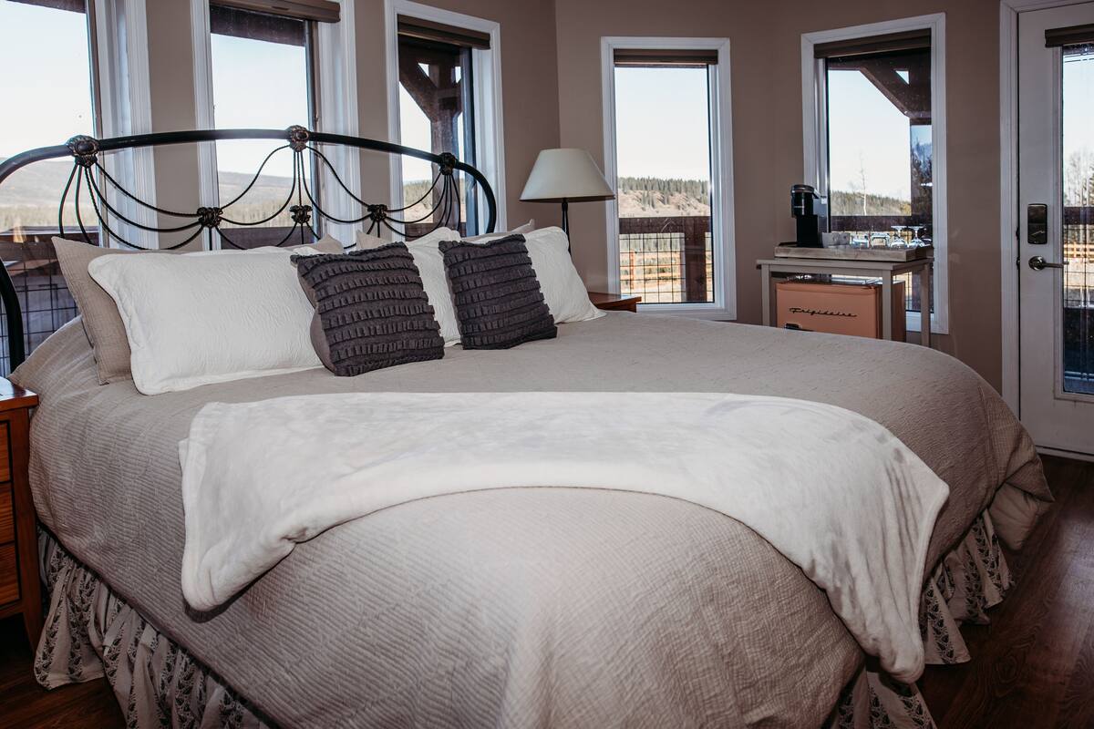 A large bed with a headboard, two bedside tables, a lamp, and a view of the outdoors through large windows.