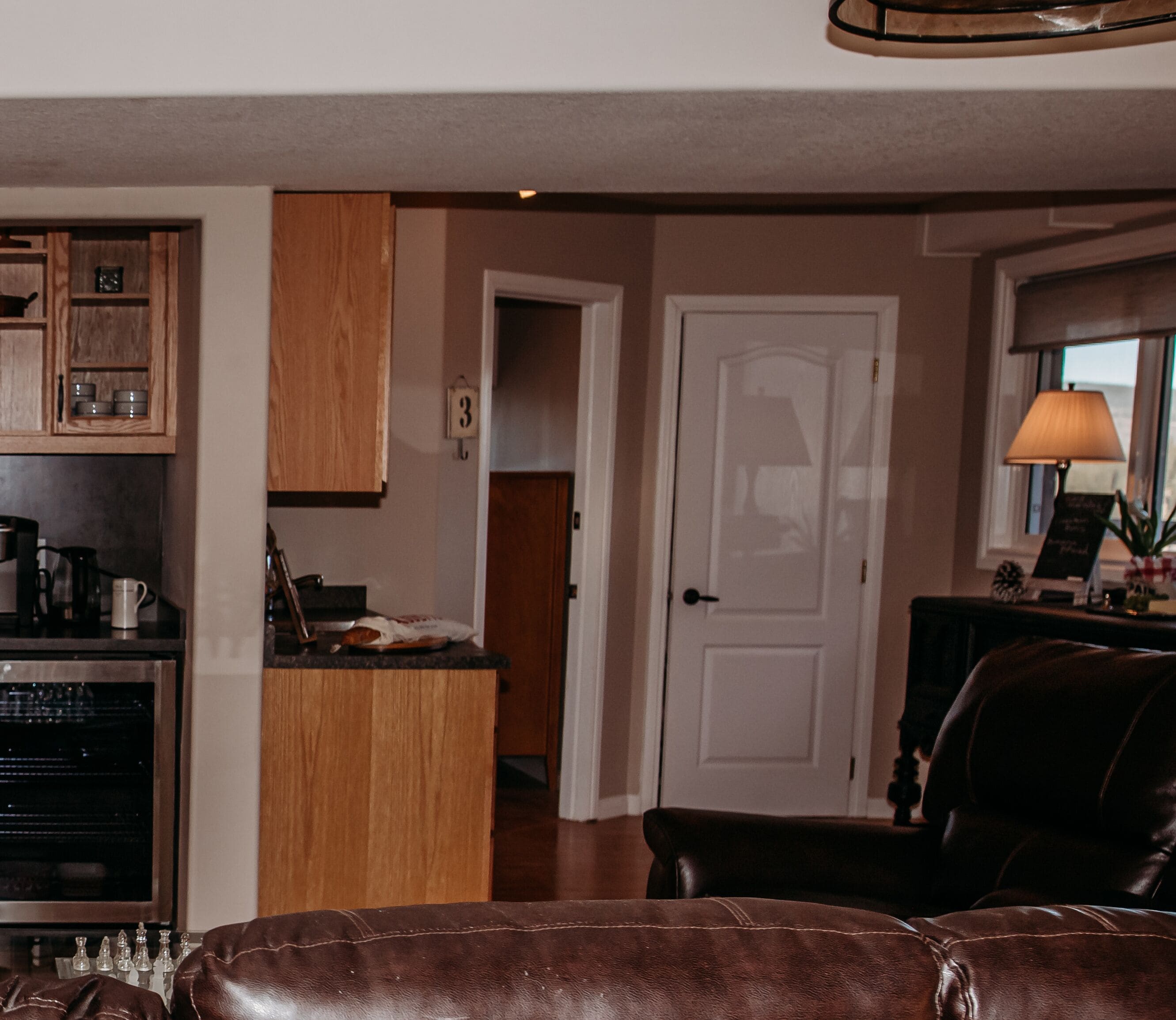 A modern kitchen with wooden cabinets, a coffee machine, and a small island. There is a door leading to another room and a lamp on a side table.