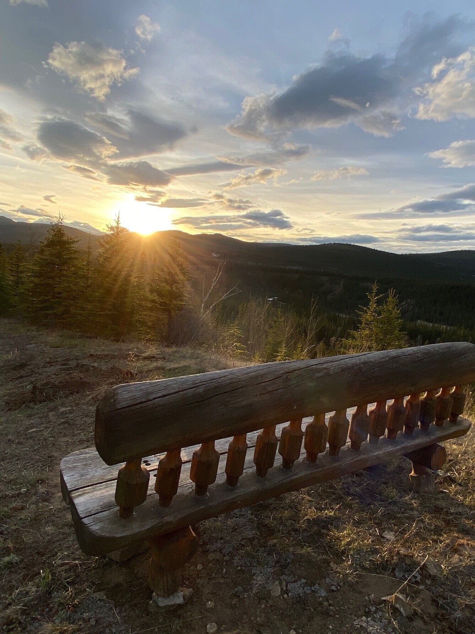 Un banco de madera con barandillas metálicas, ubicado en un sendero de tierra con vistas a un paisaje boscoso y colinas distantes bajo un cielo parcialmente nublado.