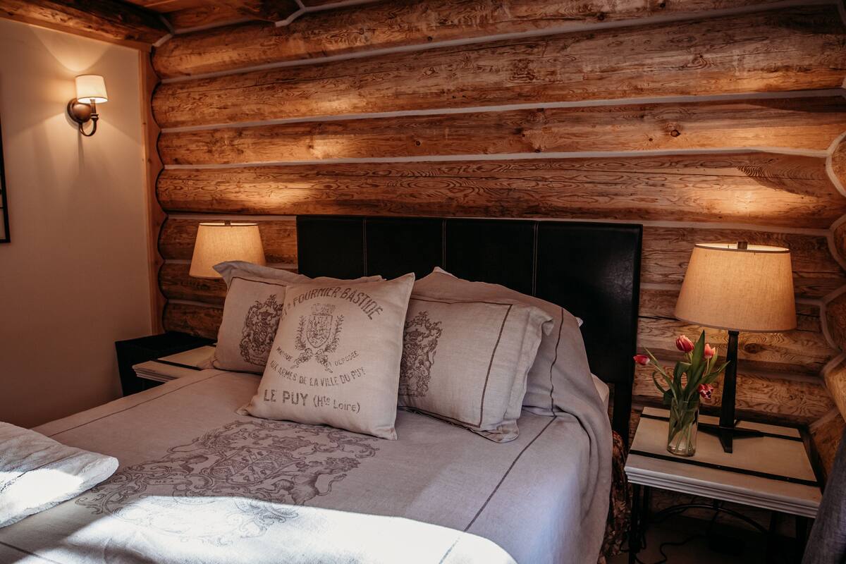 A log cabin-style bedroom with a bed, two bedside lamps, a nightstand, and a vase with flowers.