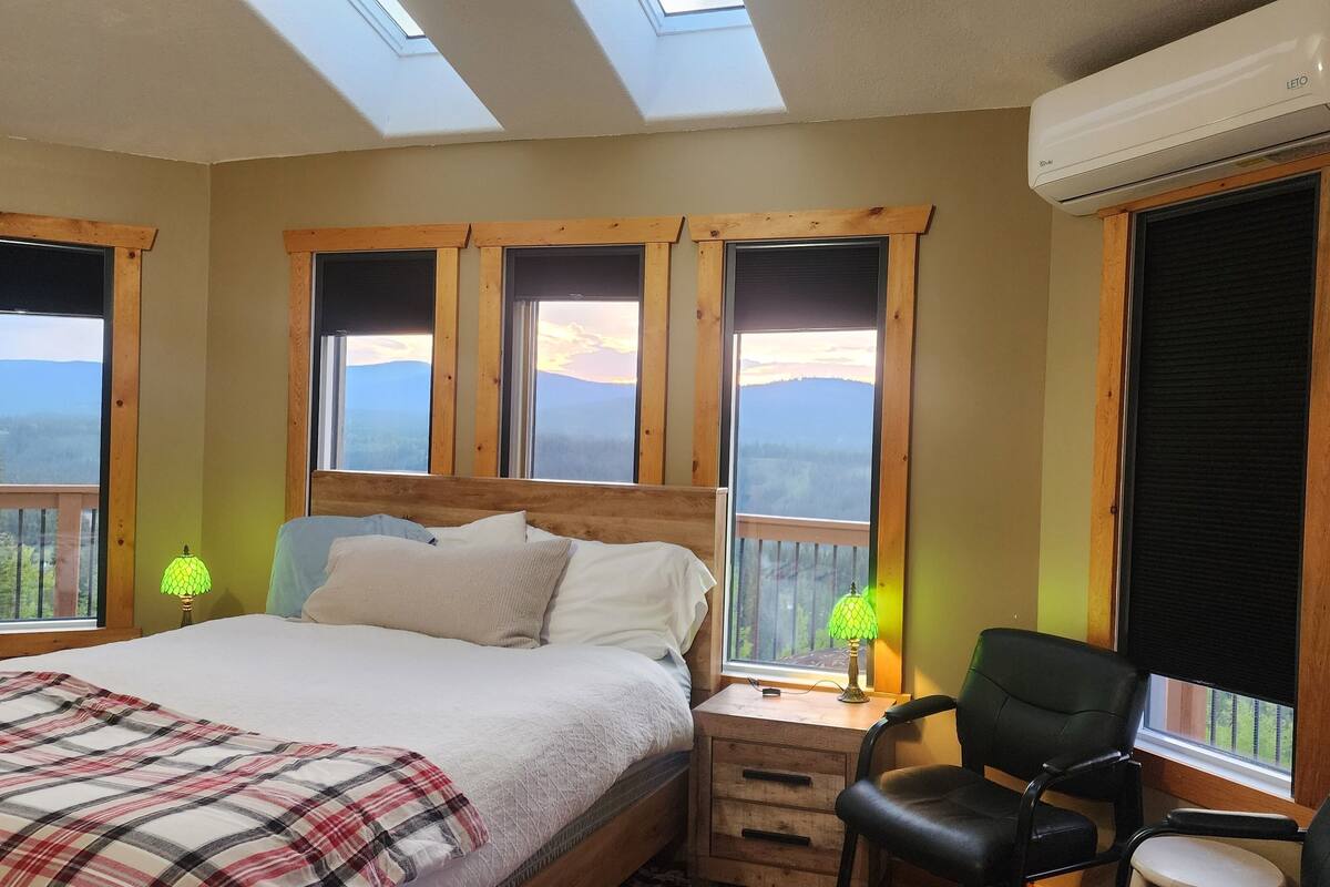 A bedroom with a bed, a desk, and a chair. There are skylights and windows with a view of mountains.