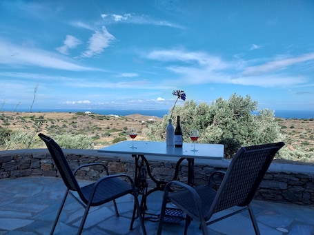 A table with two chairs and a wine bottle, overlooking a scenic landscape with olive trees and a view of the sea.