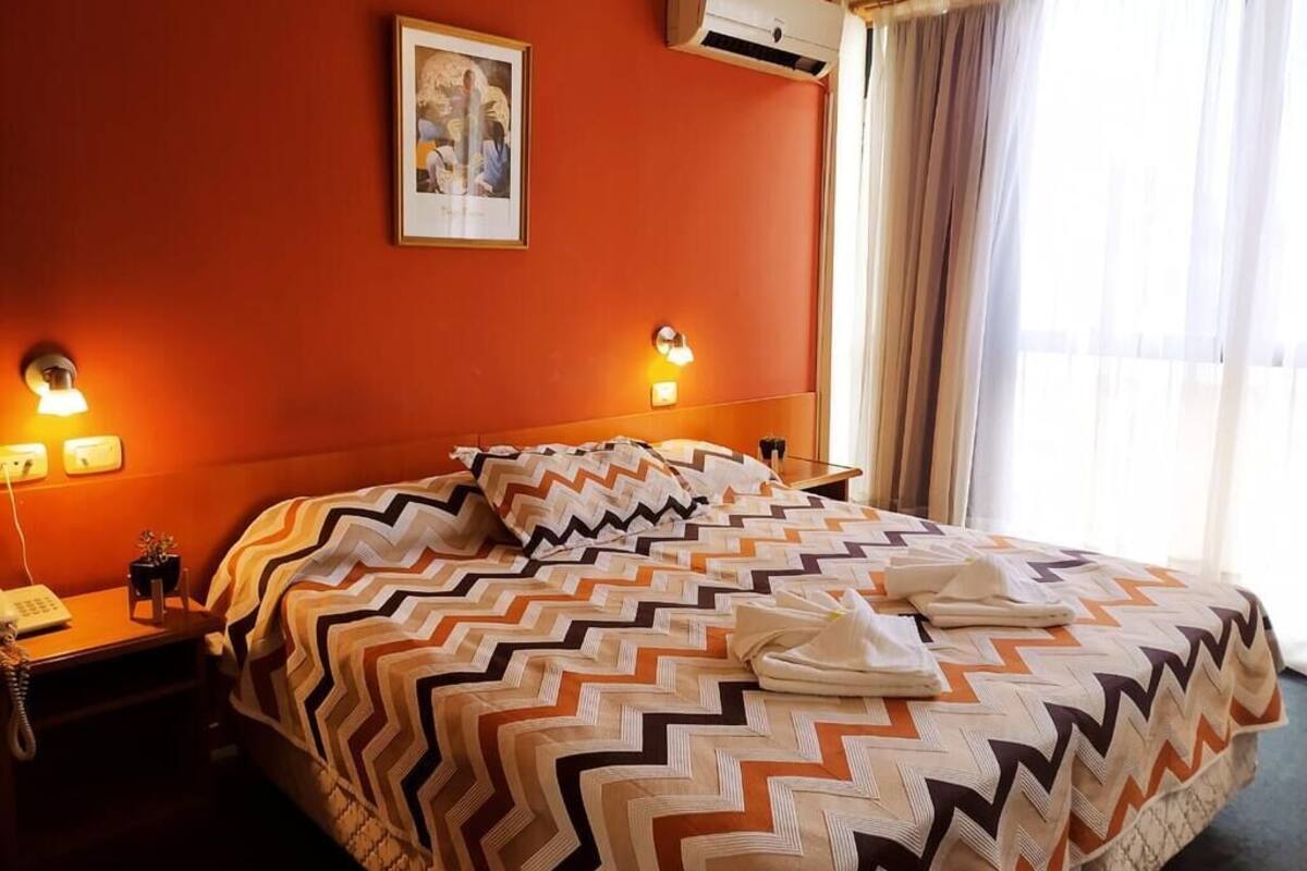 A hotel room with a large bed, two bedside lamps, an air conditioner, a framed picture on the wall, and a window with curtains.