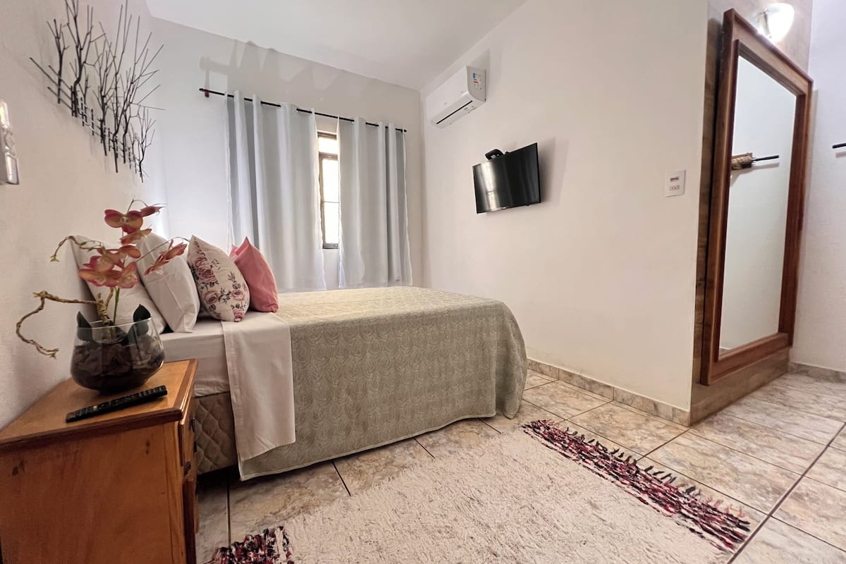 A bedroom with a bed, a TV, a window with curtains, a bedside table with a vase, and a rug.