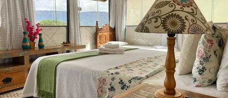 Habitación de hotel con cama, mesita de noche con lámpara, ventana con cortinas y vistas a las montañas.
