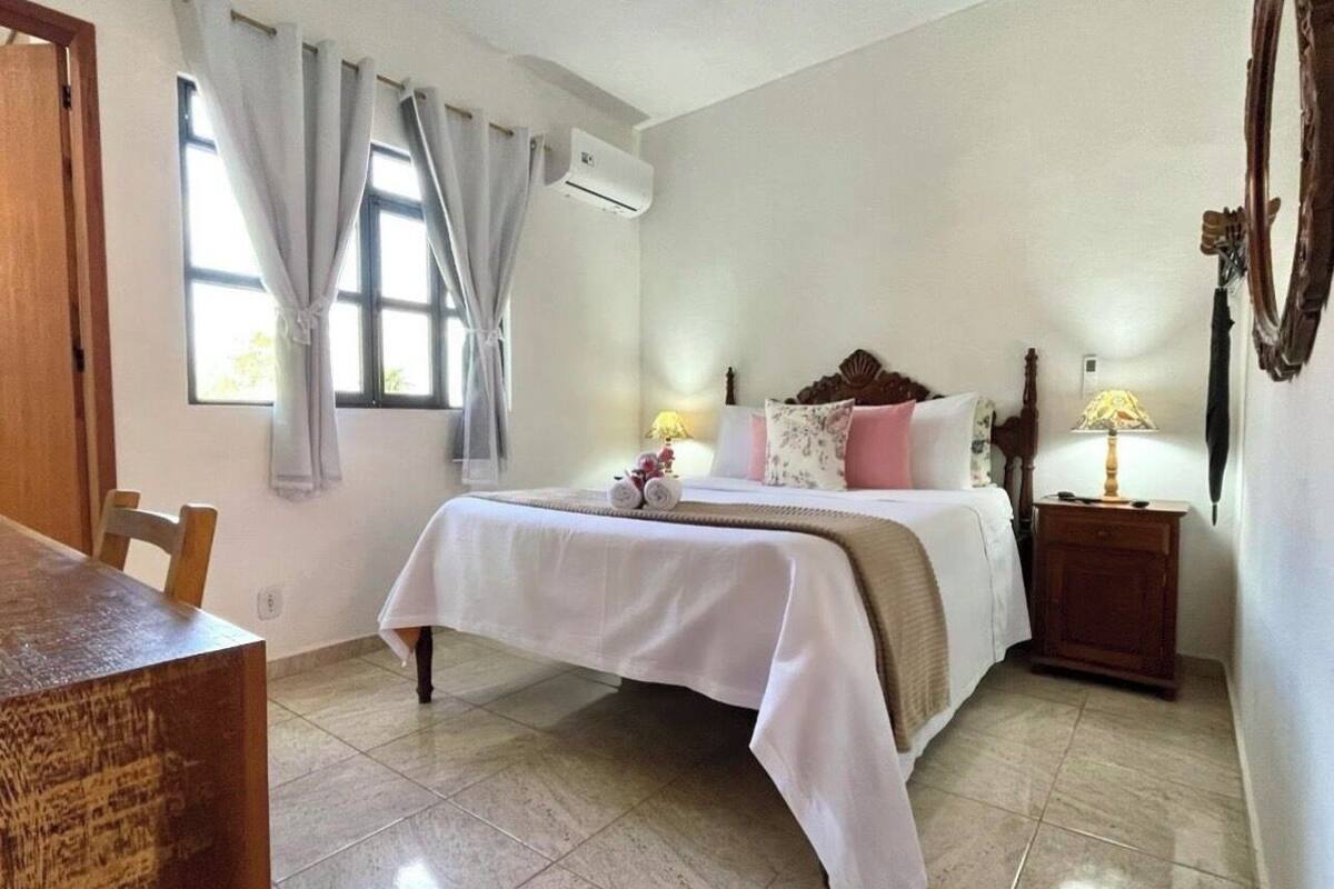 A bedroom with a bed, bedside lamps, a wooden nightstand, a window with curtains, and an air conditioning unit.
