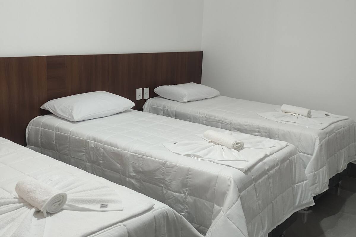 Een hotelkamer met drie eenpersoonsbedden, elk met witte lakens en kussens, en een houten hoofdbord.