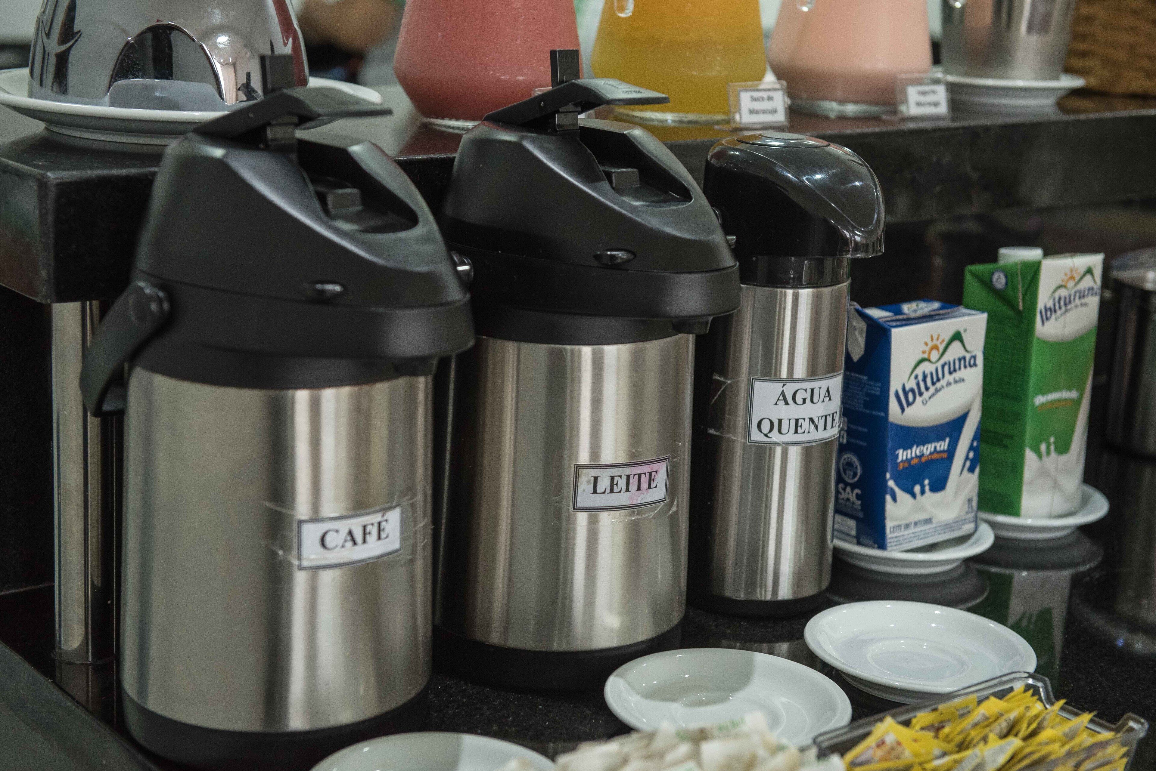 Dispensadores de bebidas de acero inoxidable con las etiquetas 'Café' y 'Leite' sobre una encimera con cartones de leche y un plato de aperitivos.