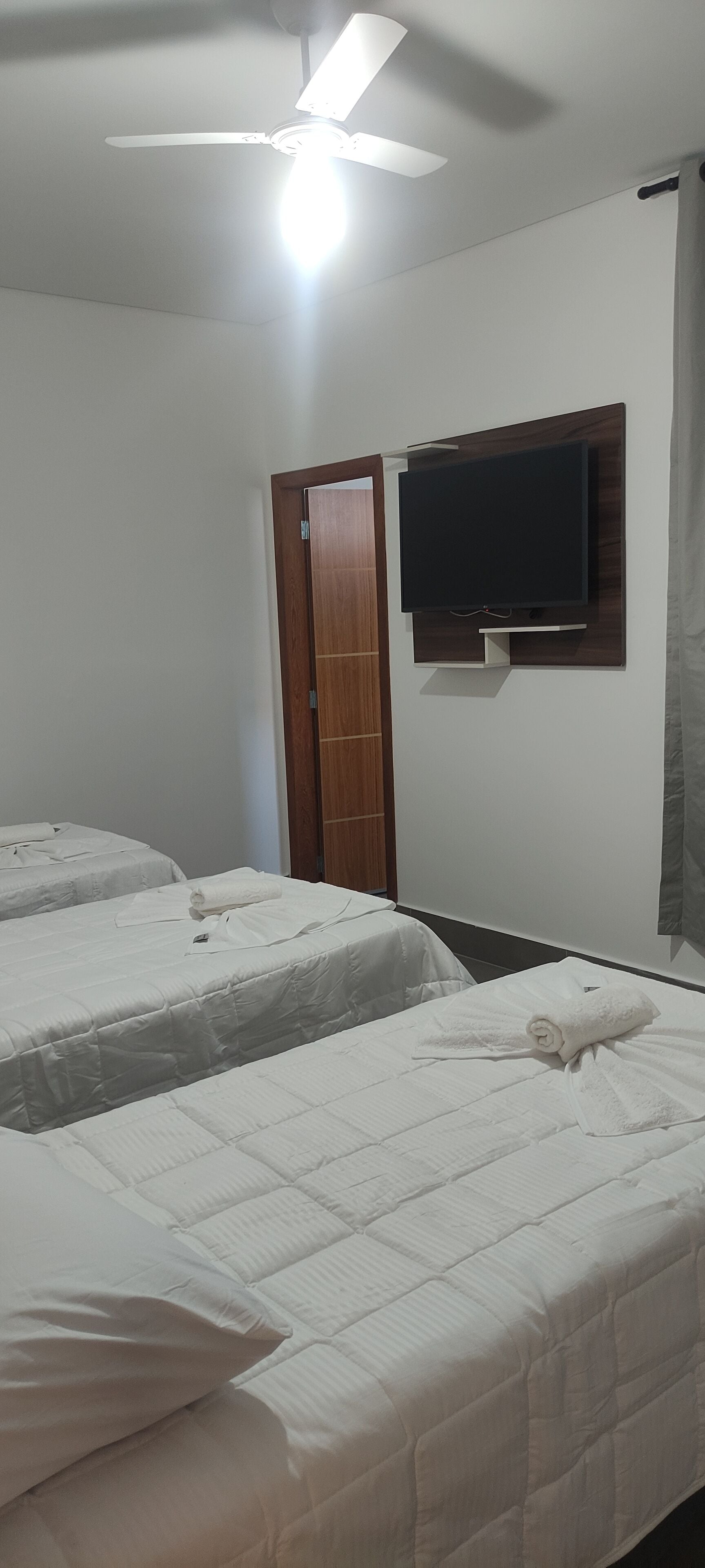 Quarto de hotel com duas camas, um ventilador de teto, uma televisão de tela plana na parede e uma porta de madeira.