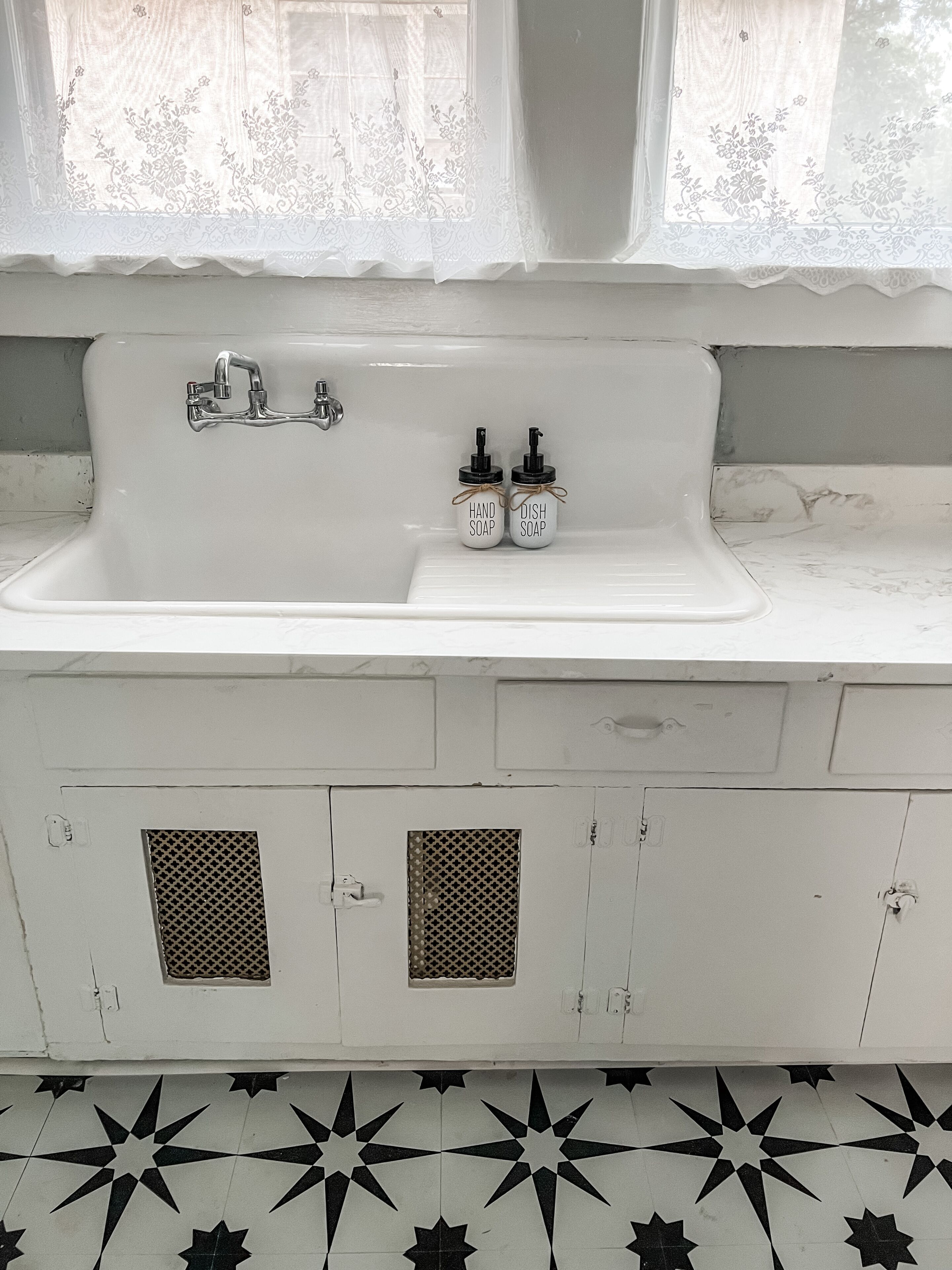 Ein Badezimmer mit einem weißen Waschbecken, zwei Seifenspendern, einem Wasserhahn und einem Fenster mit Spitzenvorhängen.