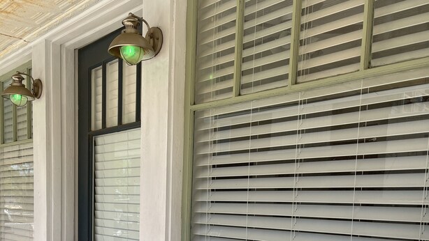 Una habitación con un ventanal con persianas, una puerta con picaporte y una pequeña mesa redonda con una silla.