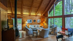 Interior acogedor de una cabaña con techo de madera, ventanas amplias, chimenea y zona de estar con sofá y sillones.